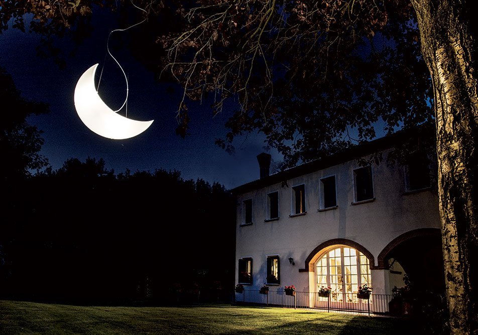 Mazzega_moon_pendant_02 | rewire Night scene of a white two-story house with a warmly lit arched doorway and windows, under a large crescent light in the sky.