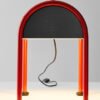 "Valigia", features a small handle and two load-bearing arches with red piping. Its metal vault houses the light source, making it a portable piece of architecture. The supporting structure and lampshade come in a red and black finish.