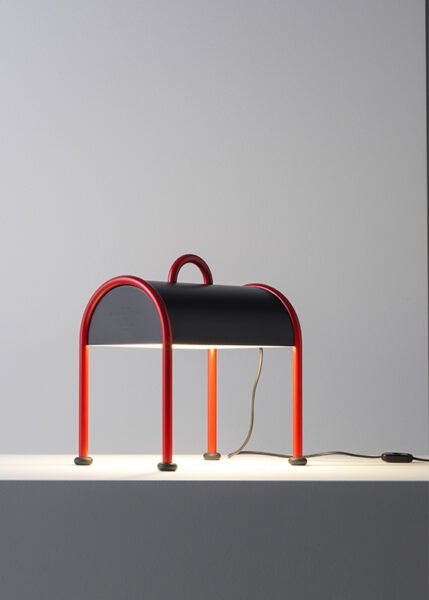 "Valigia", features a small handle and two load-bearing arches with red piping. Its metal vault houses the light source, making it a portable piece of architecture. The supporting structure and lampshade come in a red and black finish.