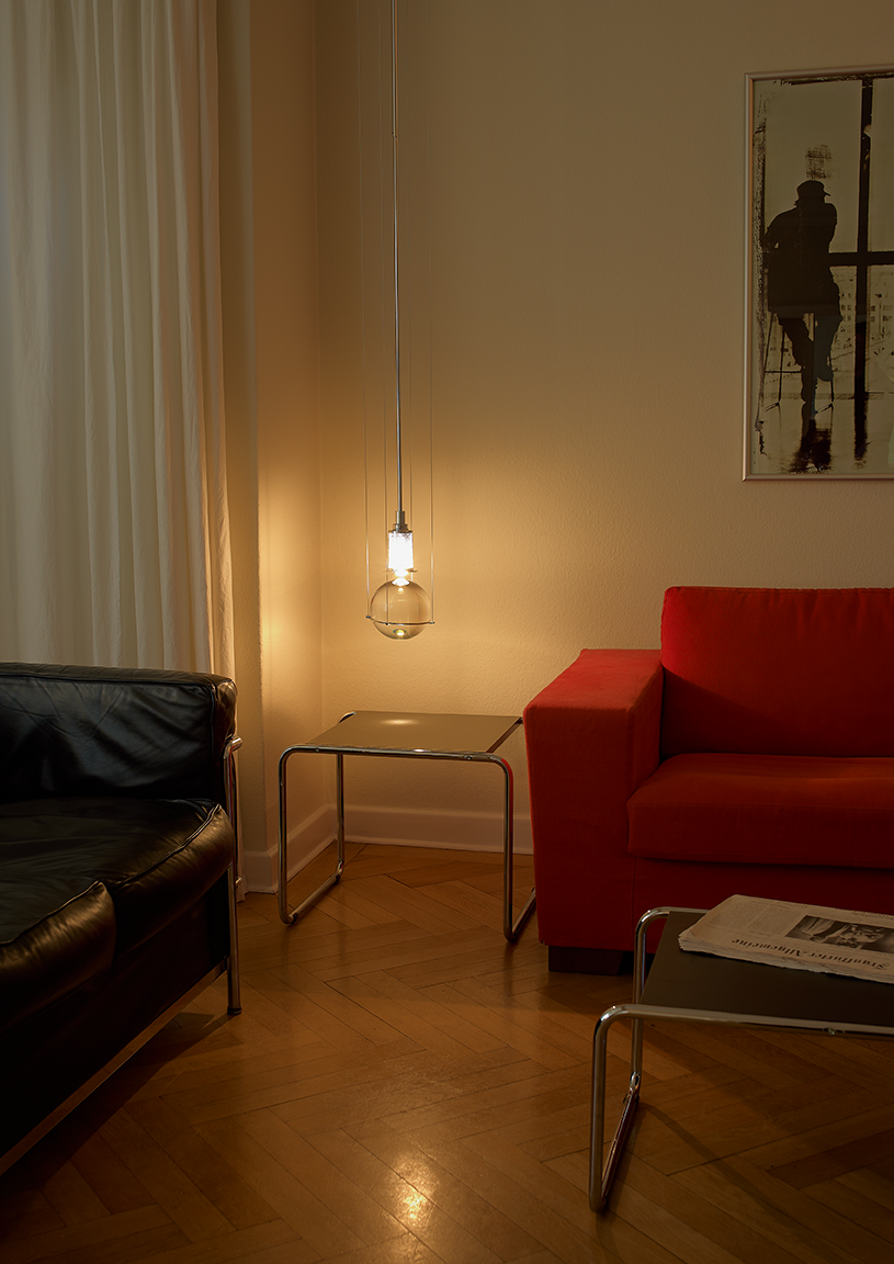 Hl3s81-ambiente_web_1 | rewire Cozy living room corner with a black leather sofa on the left and a red sofa on the right, a glass-topped metal coffee table, and warm pendant light near a framed wall photo.