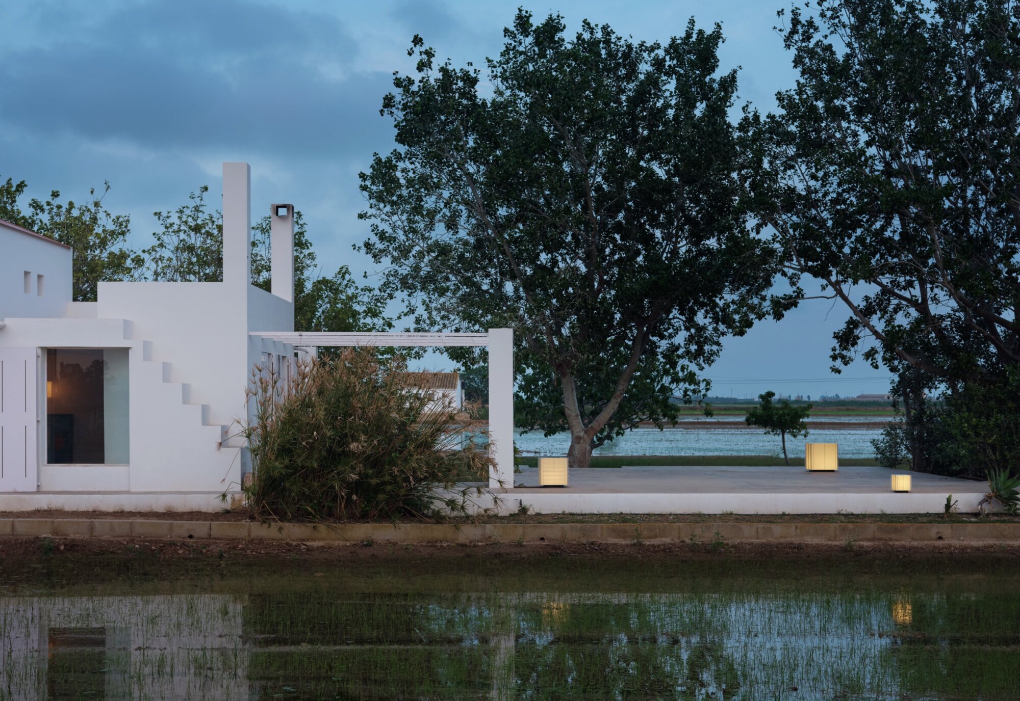 Screenshot | rewire White modern house with stepped exterior walls and terraces beside a calm water feature, under a blue dusk sky with trees nearby.