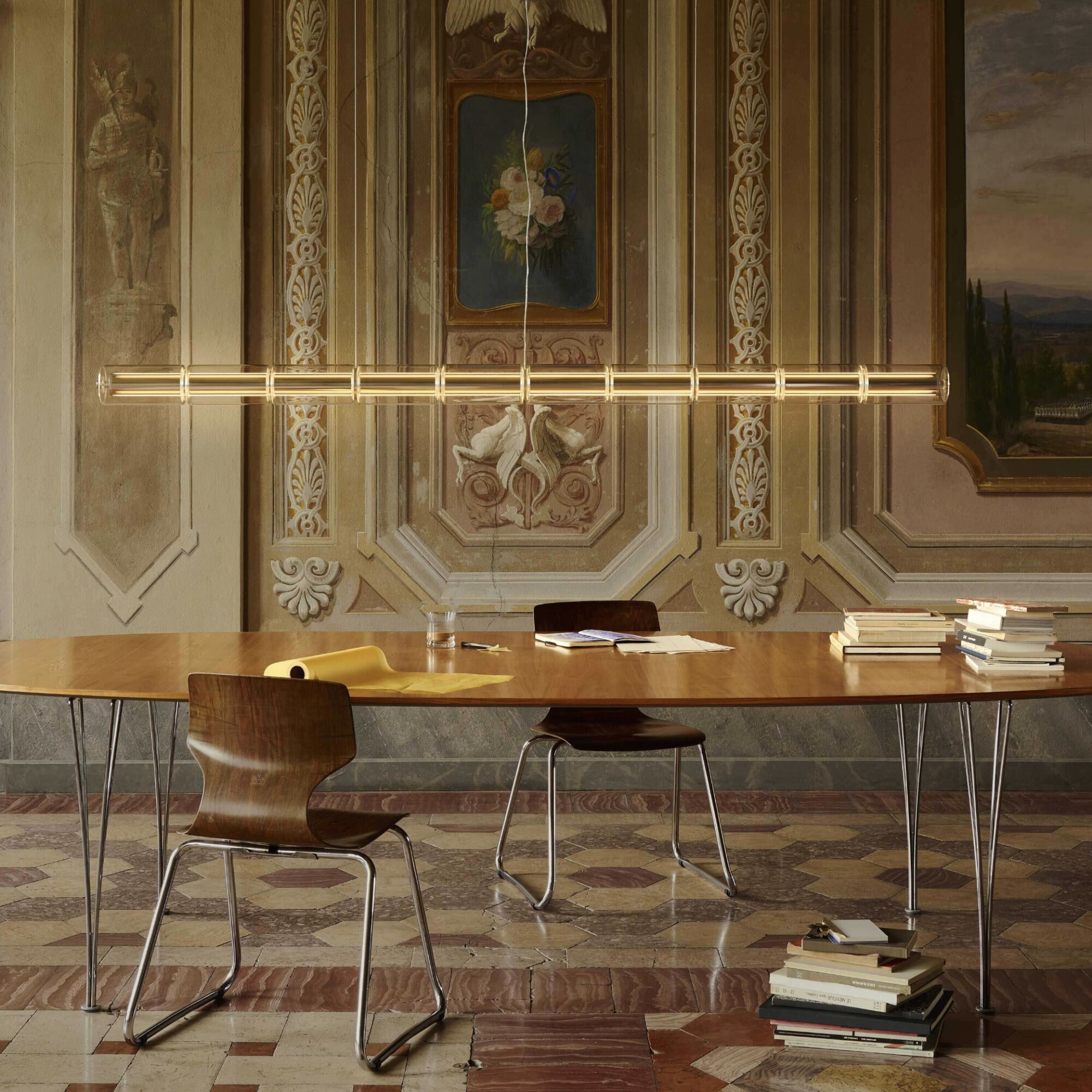 Screenshot | rewire Empty meeting room with a long wooden table, scattered papers, and stacks of books against a richly ornamented wall backdrop.