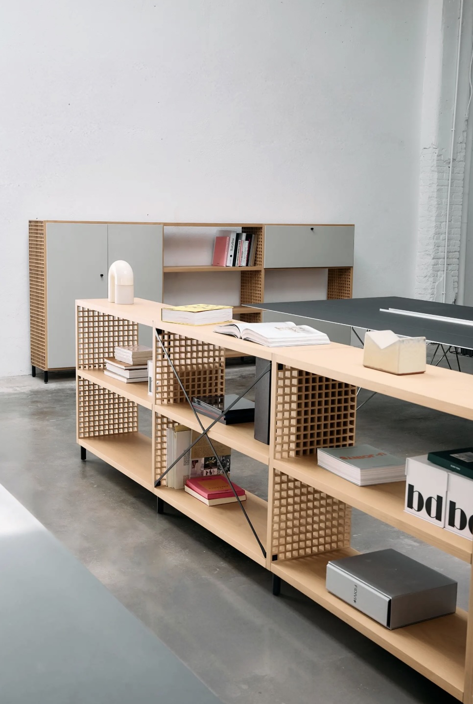 Screenshot | rewire Open-workspace scene with a long wooden table, lattice-panel shelving, and books; gray storage cabinets against a white wall in the background.