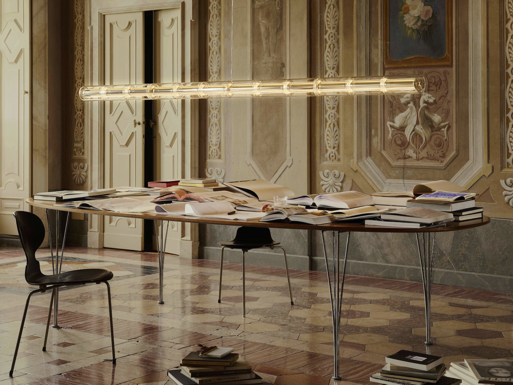 Screenshot | rewire Table laden with open books and magazines in a grand, ornate room with decorative walls and a tubular glow light fixture above.