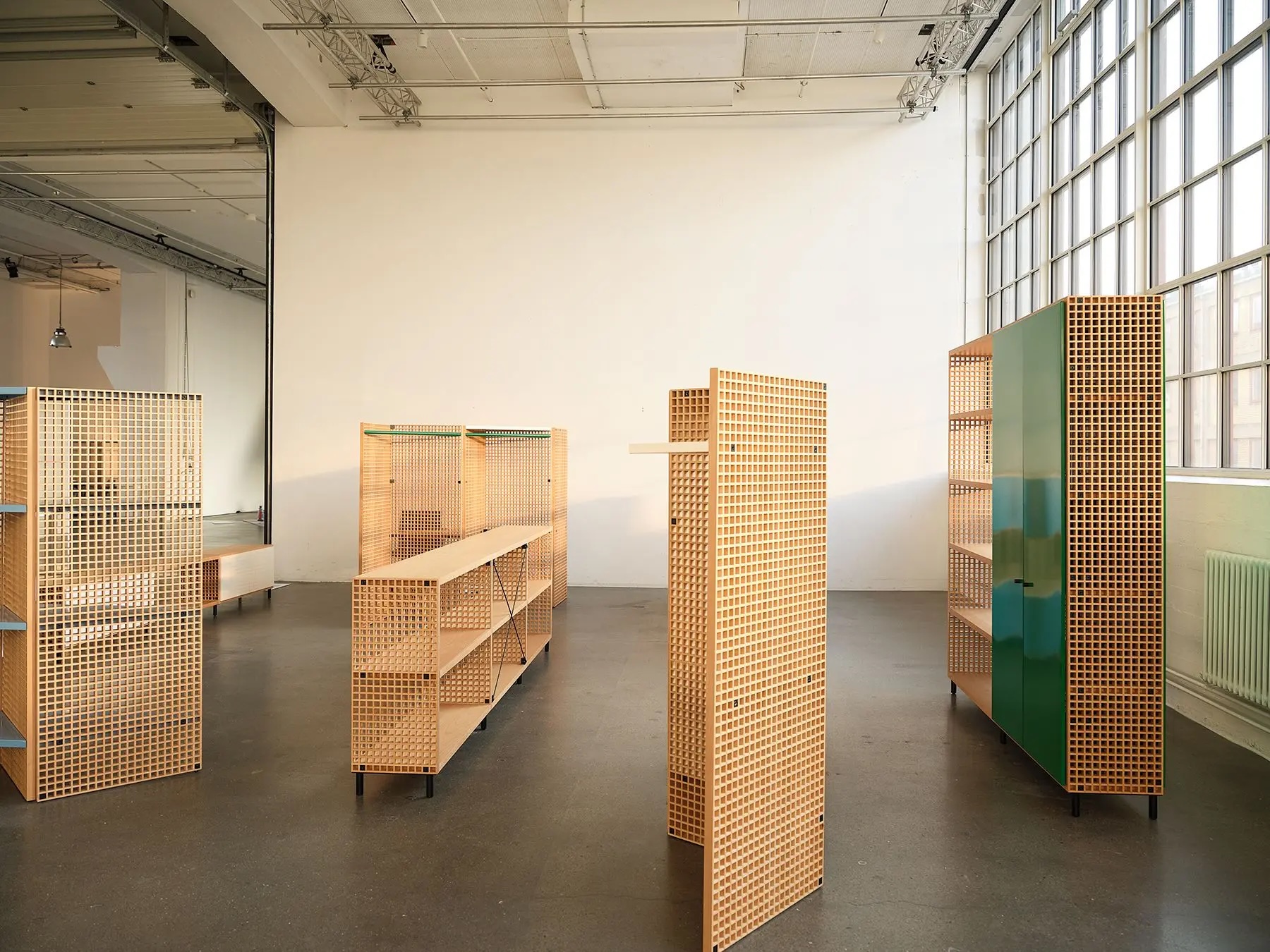 3 | rewire Industrial showroom with wooden grid shelving units arranged in rows and a tall green cabinet near large windows on the right.