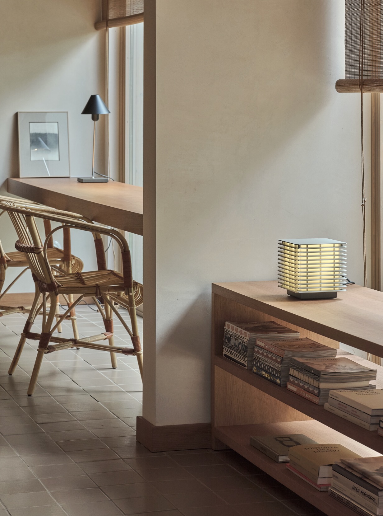 Screenshot | rewire Corner workspace with a wooden desk, black lamp, framed photo, and rattan chairs by a window in warm light.