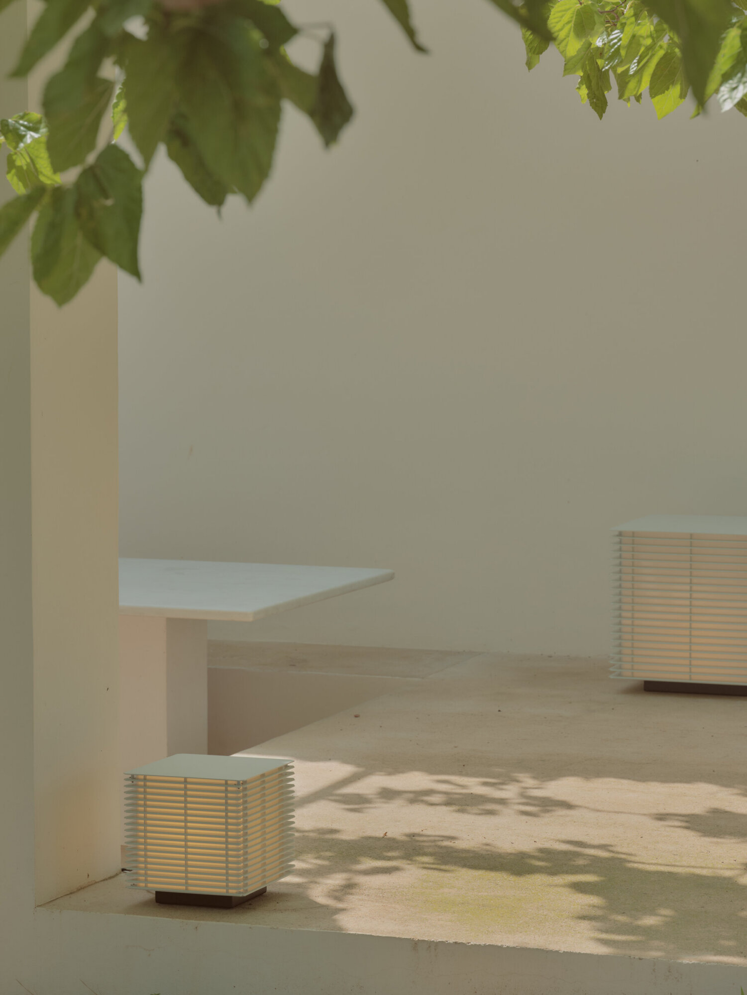 7 | rewire Minimalist outdoor courtyard with white benches, slatted metal lamps, and leafy shadows on a beige wall and concrete ground; sunny day.