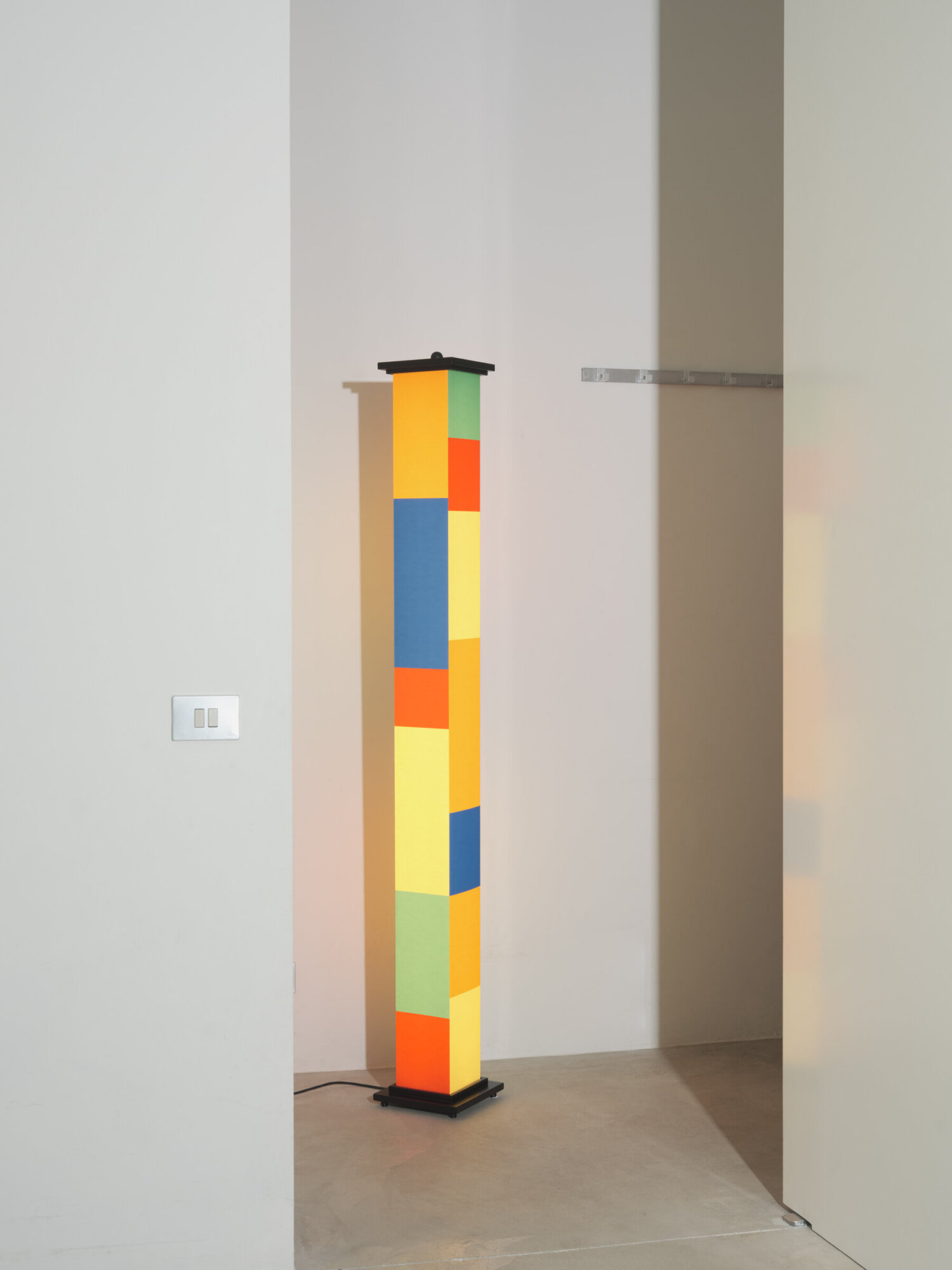 Bella-durmiente-jara-varela-santacole-2023-04-x2400 | rewire Tall vertical sculpture made of brightly colored rectangular panels stacked in yellow, blue, orange, red and green on a black base in a minimalist room.