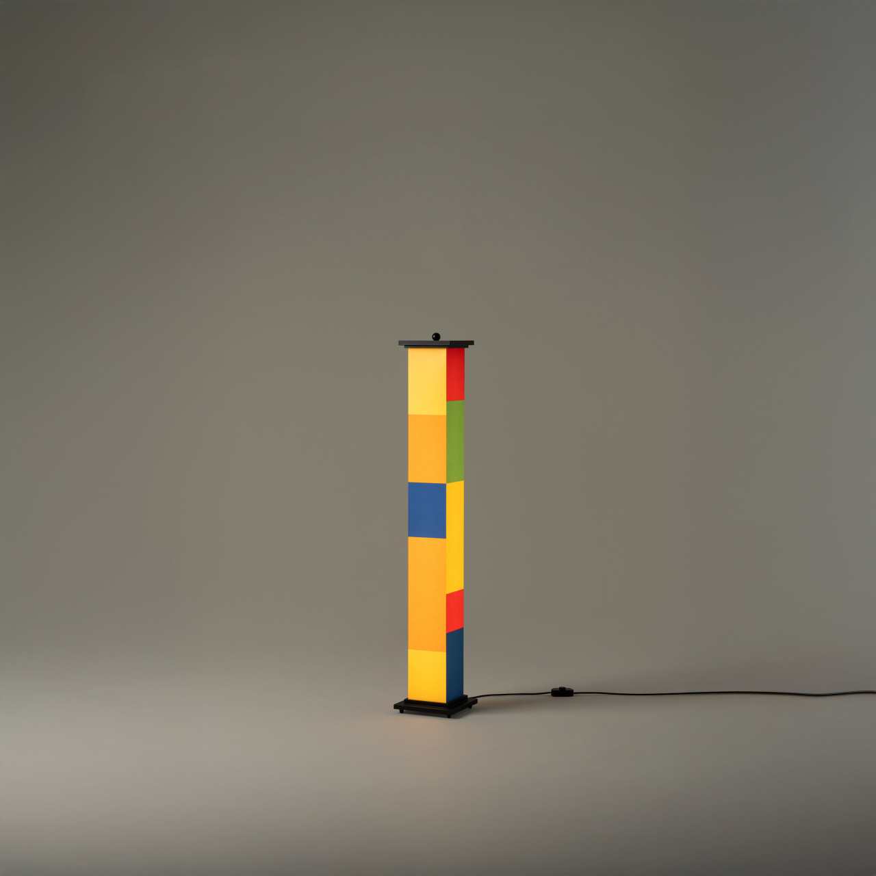 La-bella-durmiente-menor-enric-badrinas-2024-17-on-r25-x2400-1280x1280 | rewire A tall colorful striped lamp standing on a black base, illuminated against a gray backdrop with a power cord nearby.