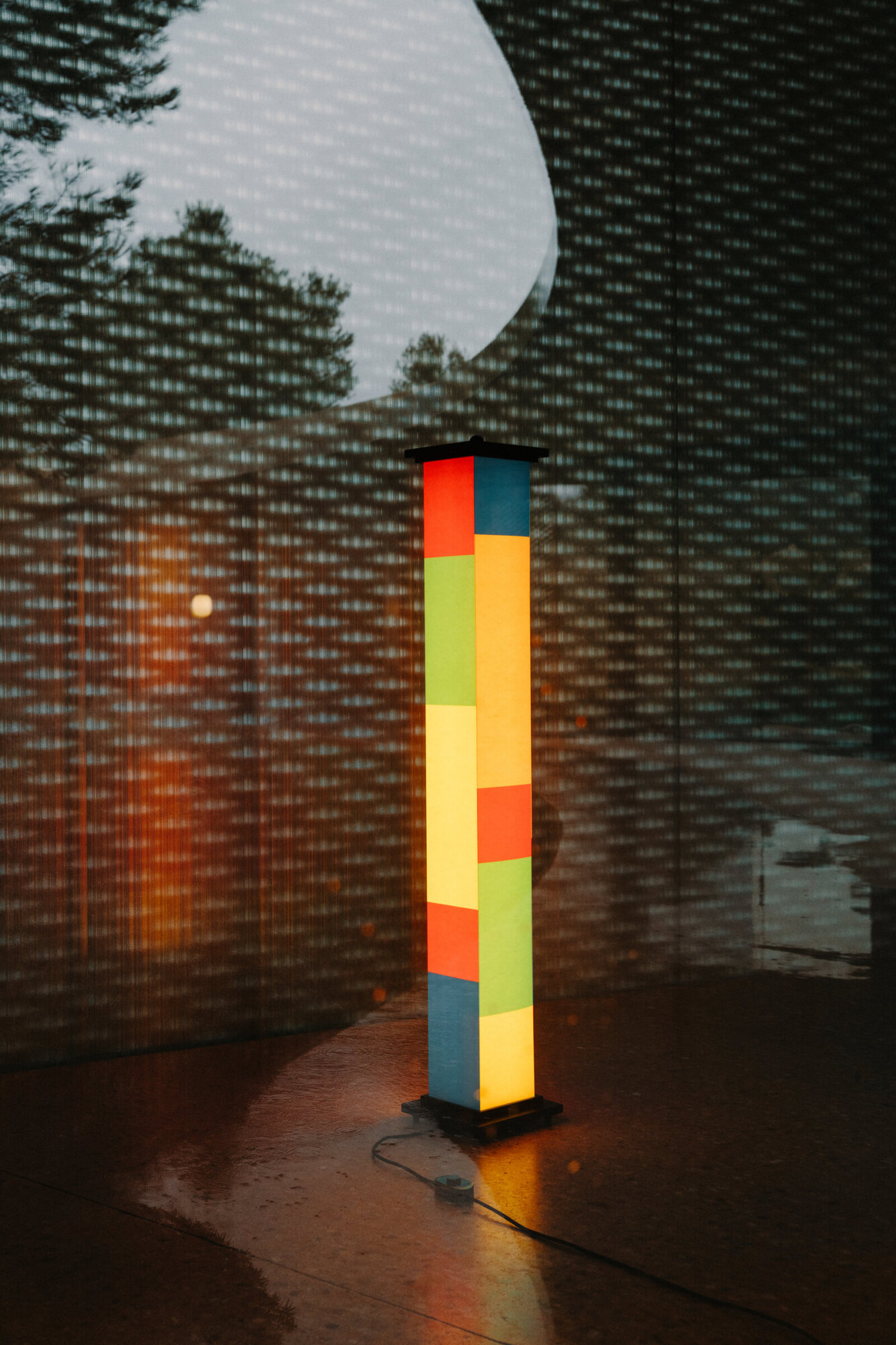 La-bella-durmiente-yosigo-2024-dsc4597-x2400 | rewire Tall pedestal sculpture made of bright color-blocked panels on a black base, illuminated in a dark room with a large white circular projection nearby.