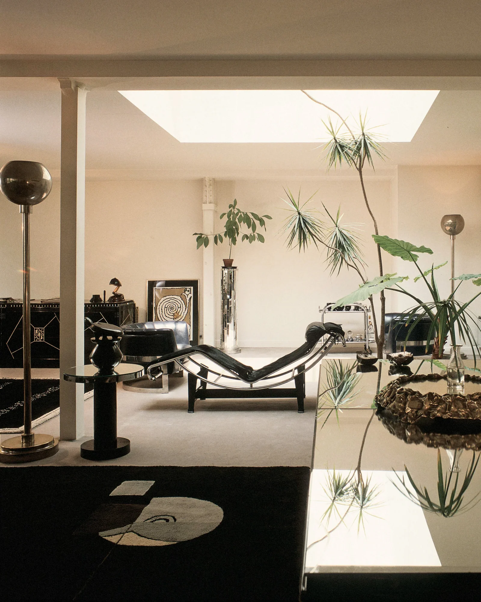 Modern living room with a black chaise lounge, glass coffee table, tall floor lamps, and large indoor plants beneath a skylight in a minimalist space.