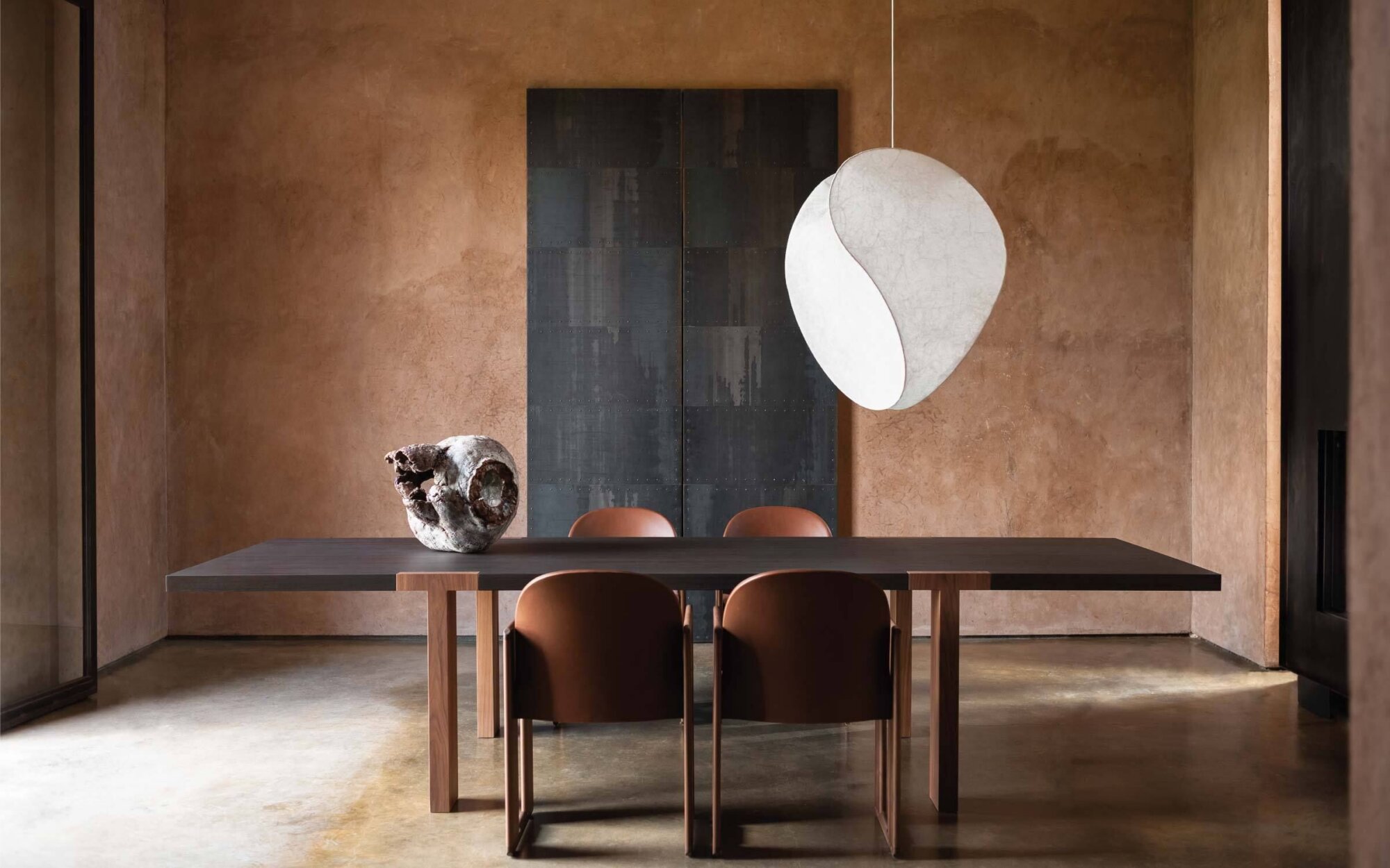 Minimalist dining room with a long wooden table, brown chairs, textured copper walls, a dark panel backdrop, and two overlapping white circular sculptures hanging above the table.