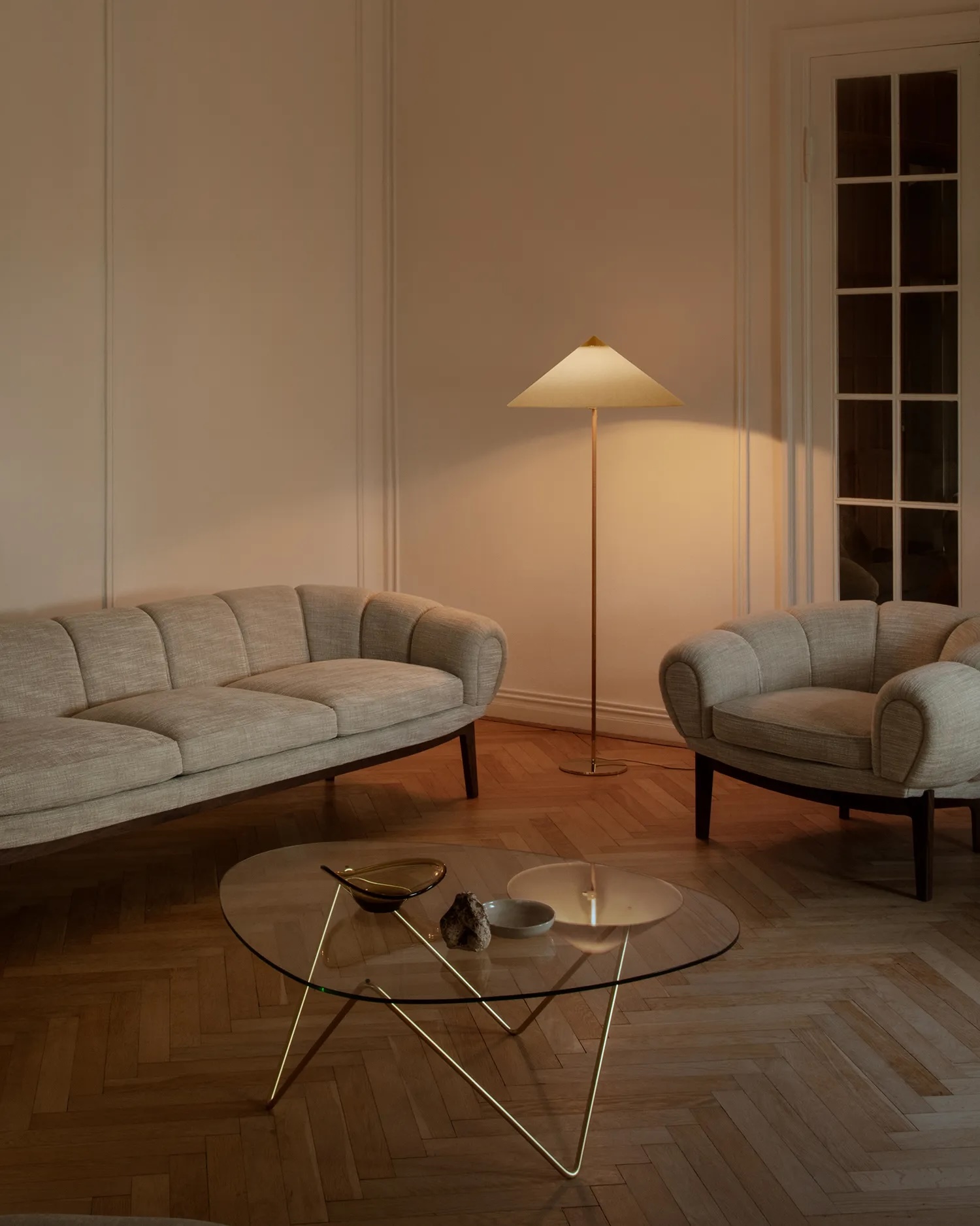 10003870_l_9602+floor+lamp_brass_canvas | rewire Living room with two light gray curved sofas, a glass coffee table, parquet floor, and a tall floor lamp casting warm light.